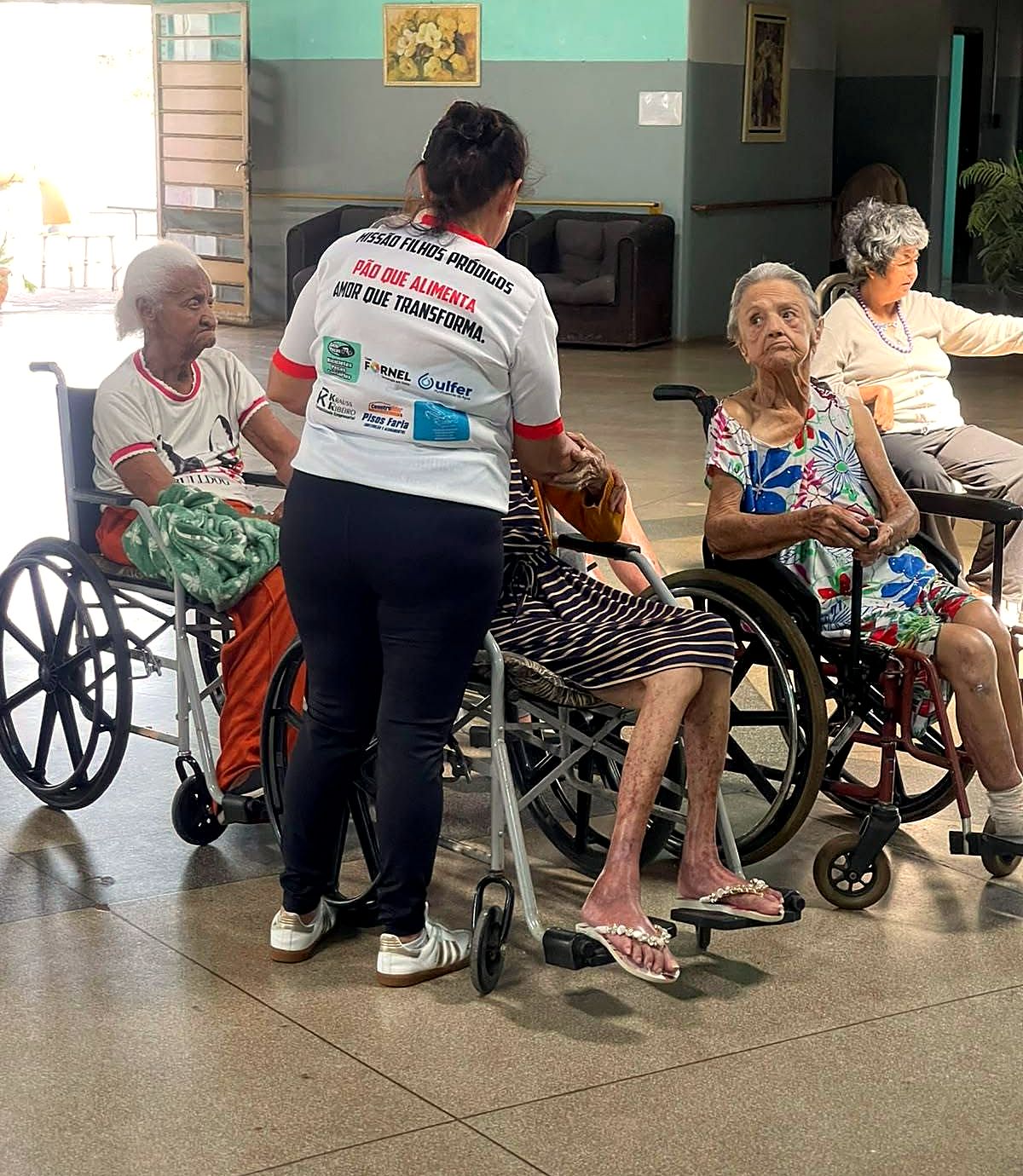 Cuidadora dando atenção a residentes em cadeira de rodas no salão da casa de repouso